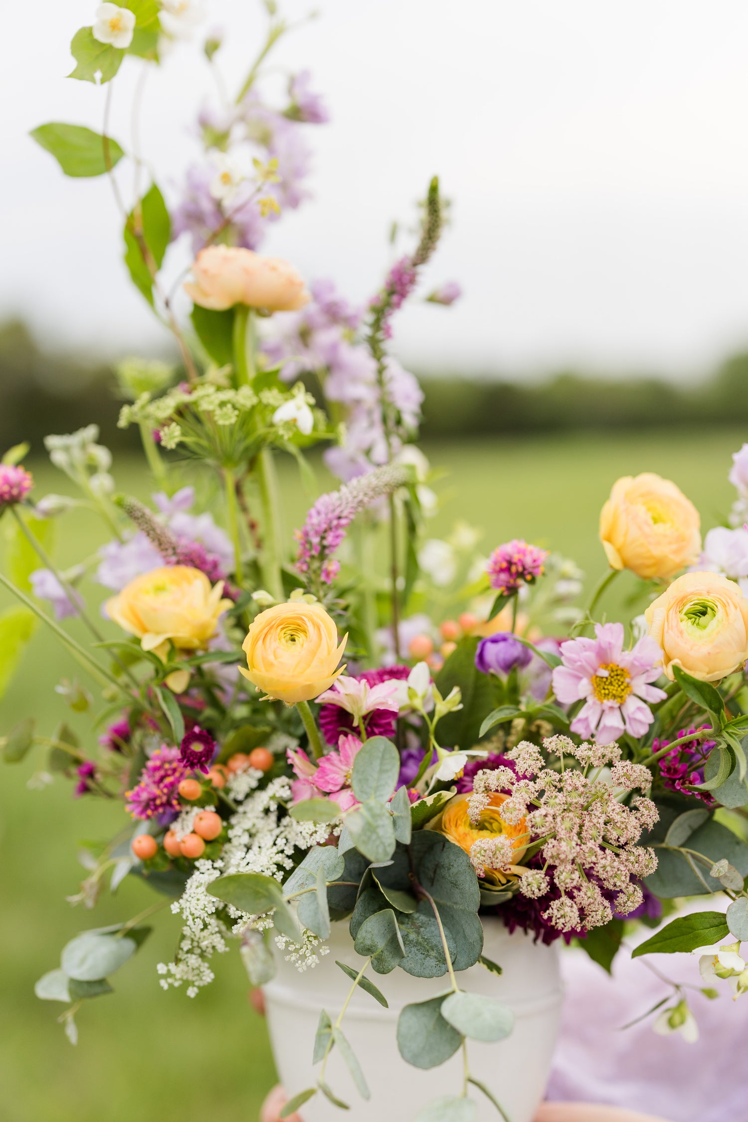 Close-up of a vibrant floral arrangement in a white vase, featuring yellow ranunculus, pink cosmos, and lush greenery in an outdoor setting.