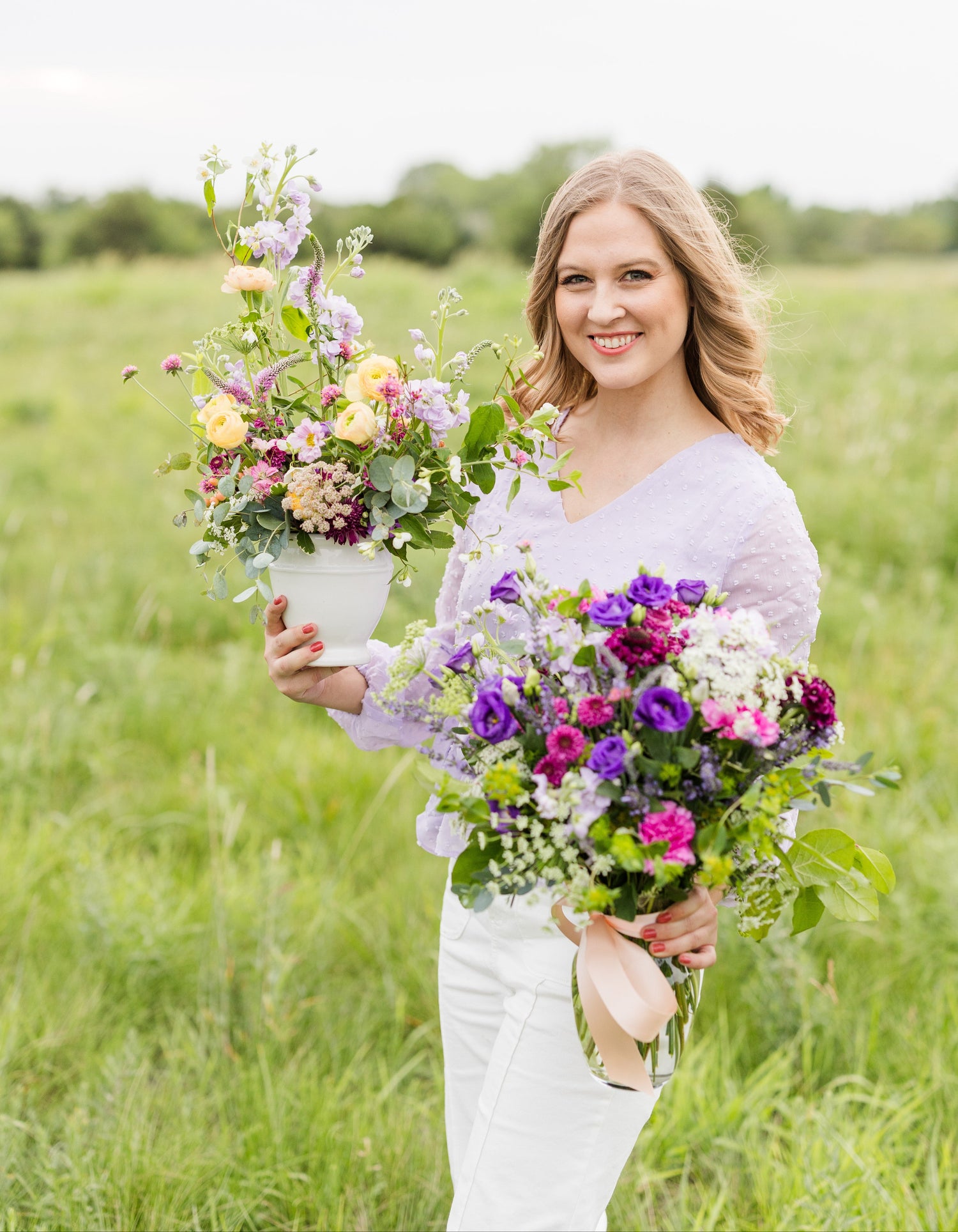 Smiling woman in a field holding two vibrant floral arrangements, one in a white vase and the other tied with a ribbon in a glass jar.